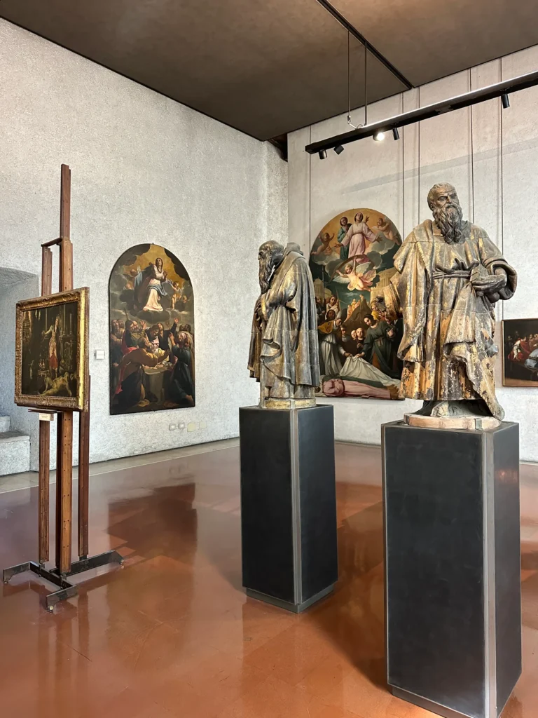 An art gallery room inside the Castelvecchio Museum in Verona, featuring two life-sized statues on pedestals and several religious-themed Renaissance paintings on the walls.