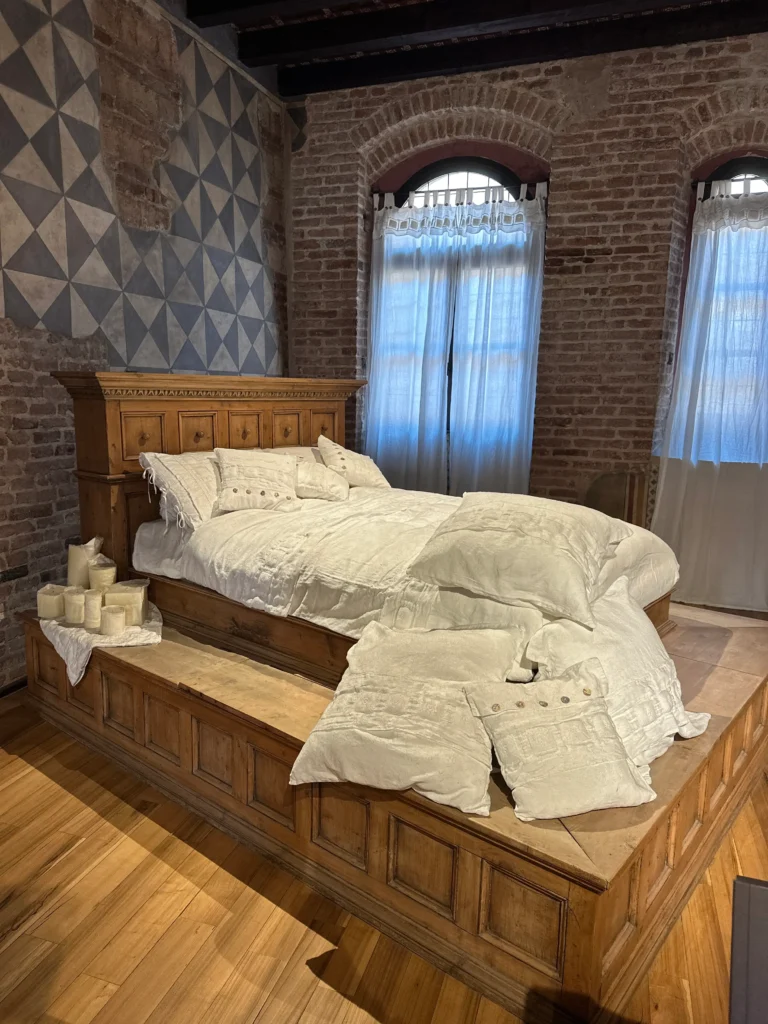 A rustic bedroom display with a carved wooden bed covered in white linens and throw pillows, set against exposed brick walls and tall windows with sheer curtains, inside Juliet’s House in Verona.
