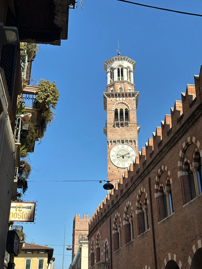 A view of the Torre dei Lamberti clock tower rising above red-brick Gothic buildings and narrow streets in Verona, under a bright blue sky.