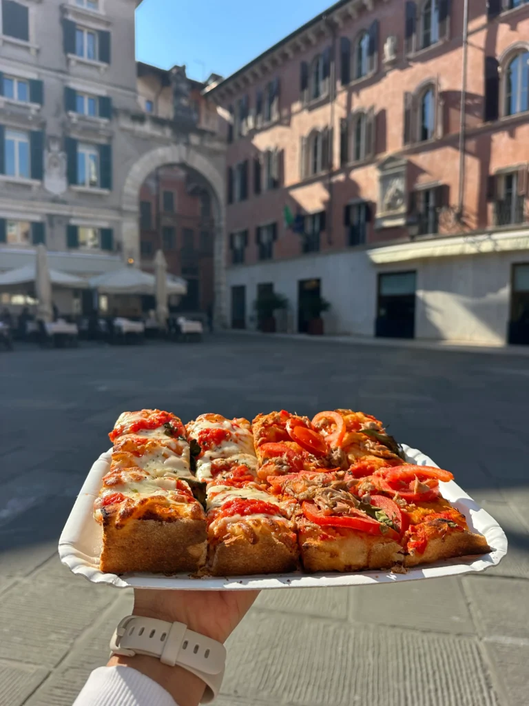 A hand holds a plate of thick, square-cut pizza from Cruncheria Verona slices topped with melted cheese, tomatoes, and herbs, in front of historic buildings and an arched entryway in a sunny piazza in Verona.