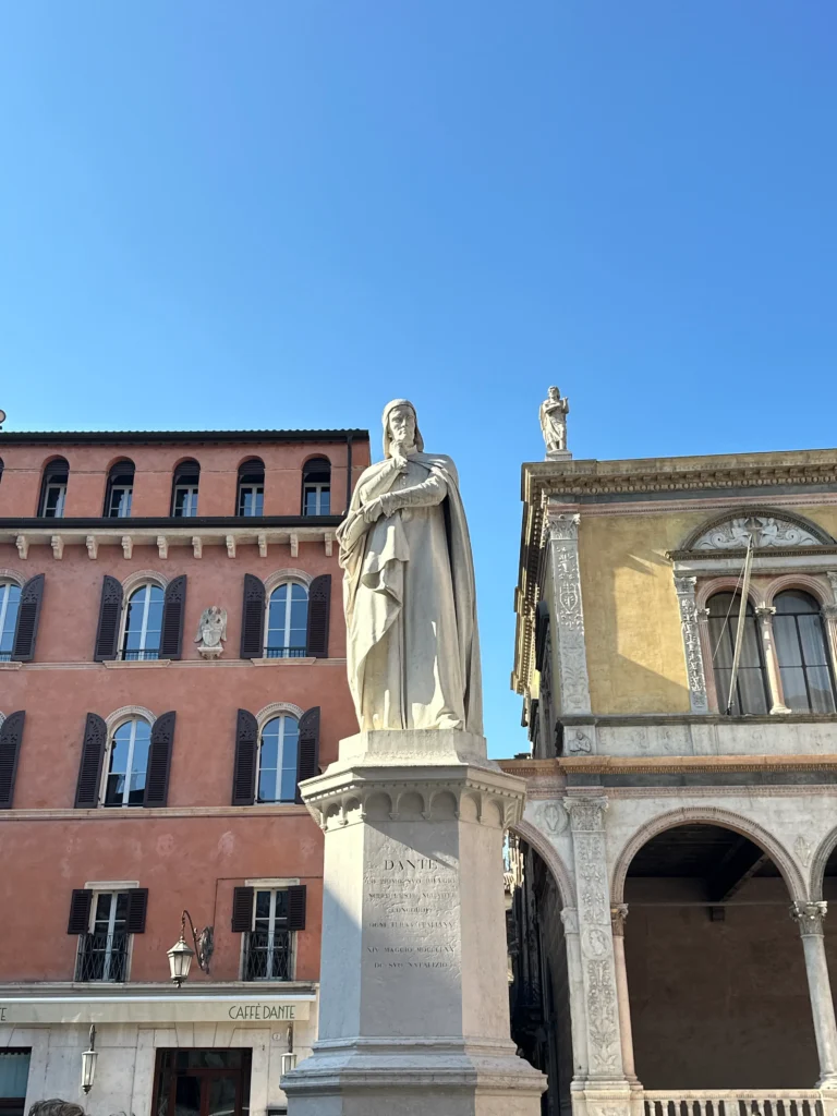 A white marble statue of Dante Alighieri standing on a pedestal in Verona’s Piazza dei Signori, with historic buildings and a café in the background under bright blue skies.