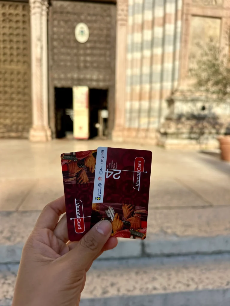 IA hand holds two red Verona Cards in front of the entrance to Verona Cathedral. A contemplation for "Is Verona Card worth it"