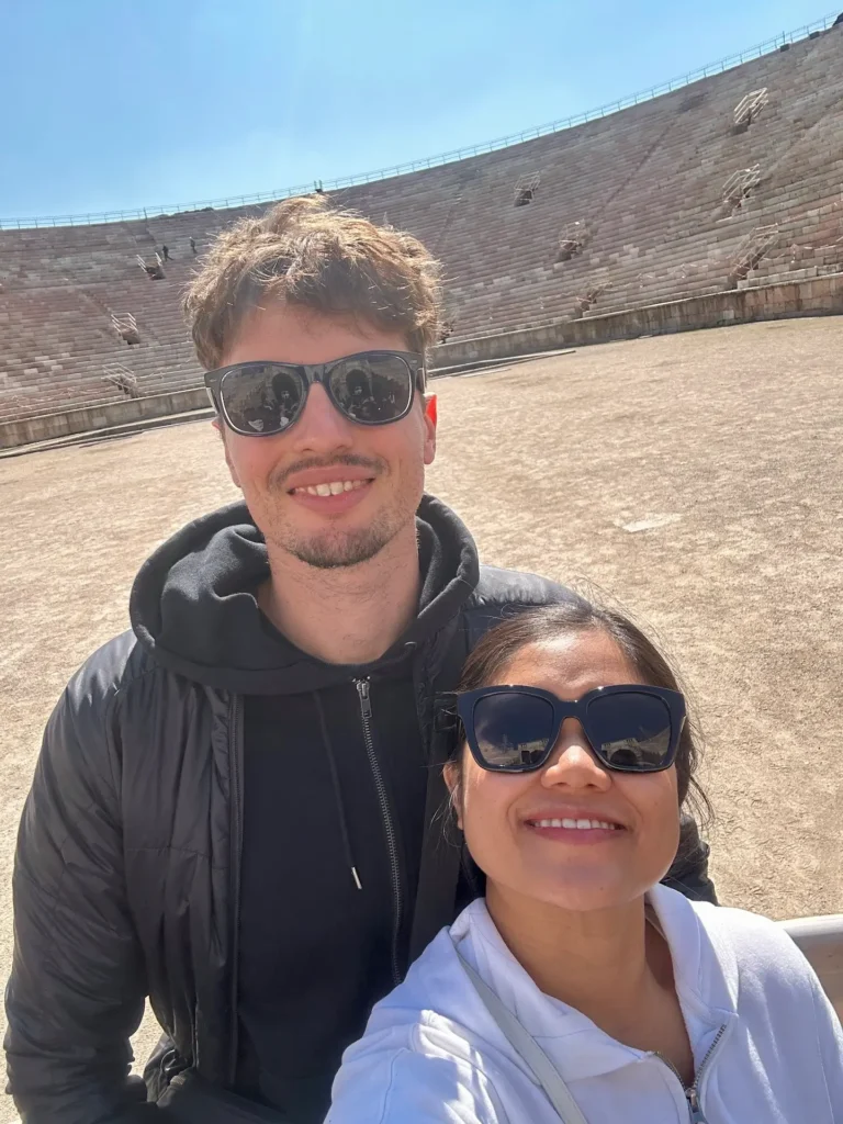Couple taking a selfie inside the Arena di Verona amphitheater with the large arena floor and stone seating behind them.
