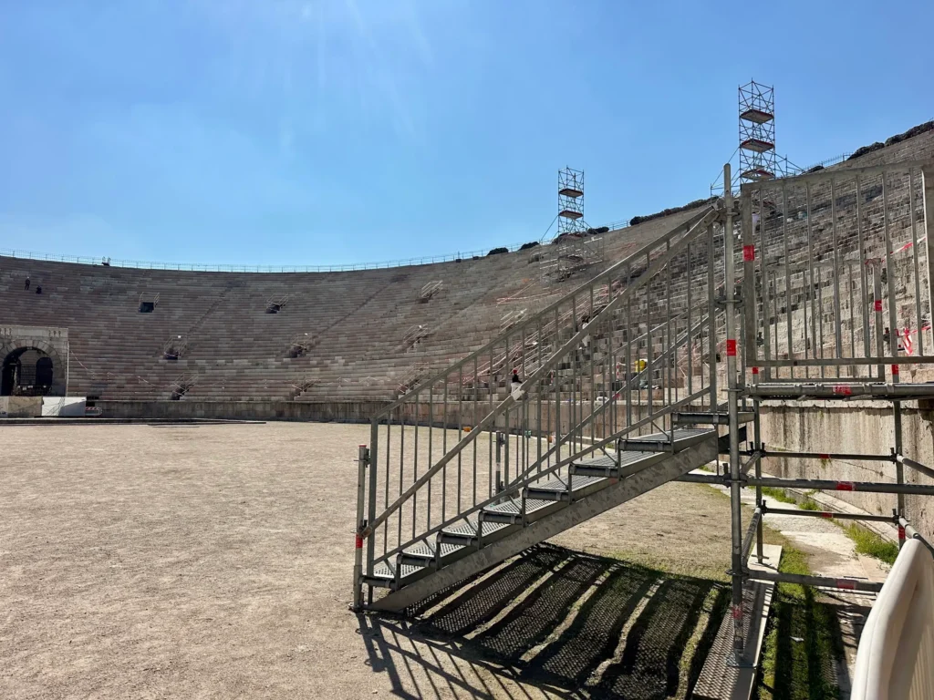 Wide interior view of the Arena di Verona showing the arena floor, surrounding stone seating, and temporary metal stairs used during renovation.