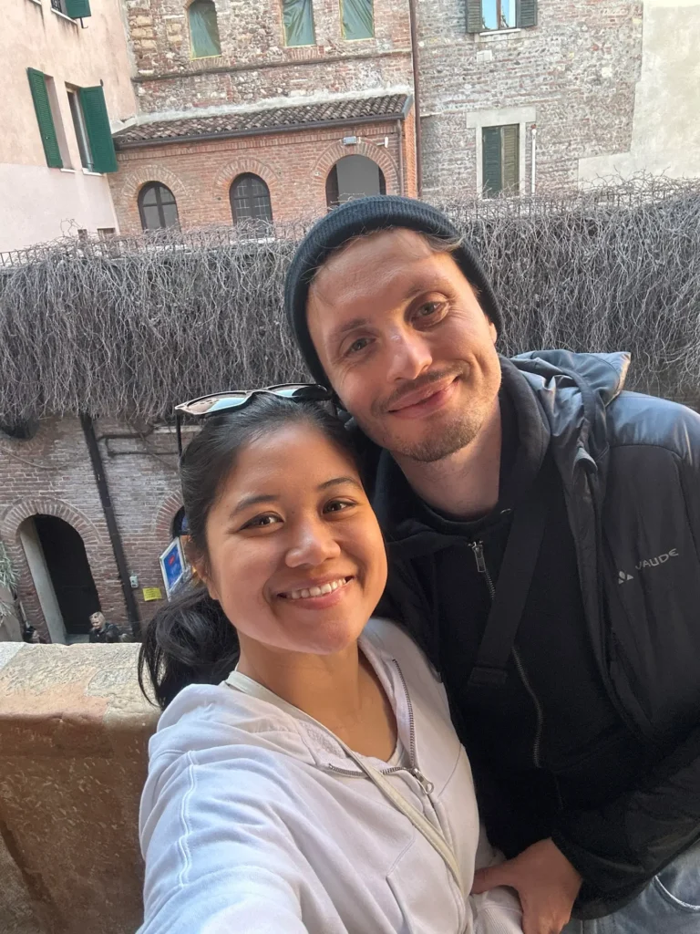 Couple taking a selfie in a courtyard at Juliet’s House in Verona, with brick walls and arched windows in the background
