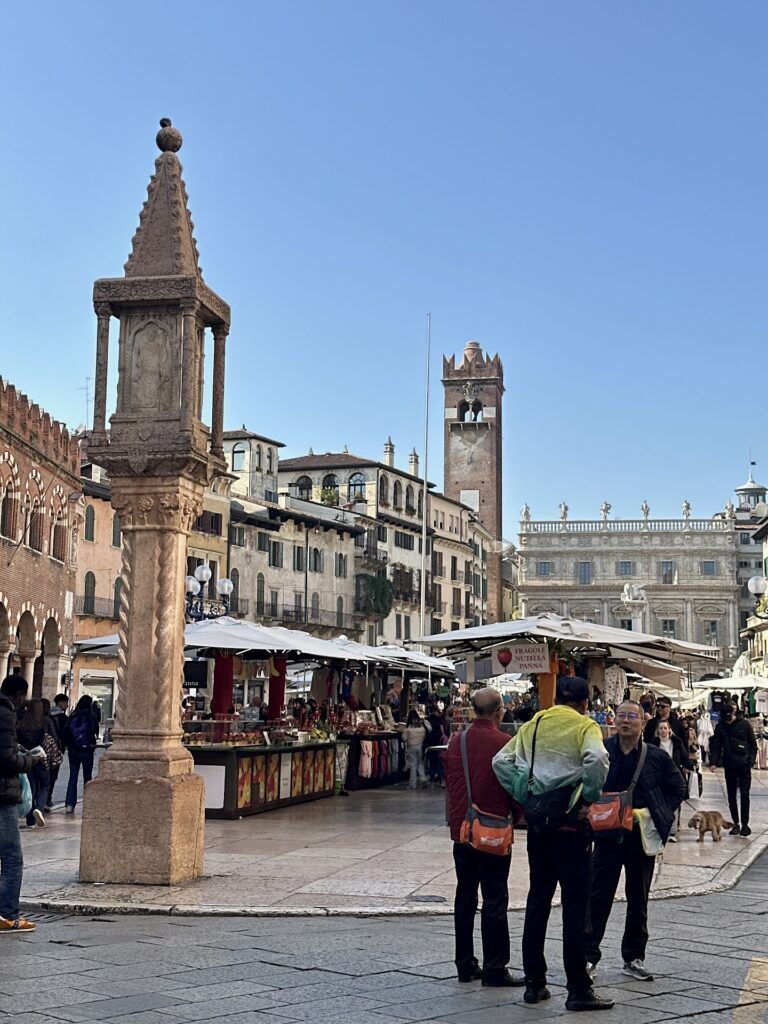 Market stalls and historic buildings in Piazza delle Erbe in Verona, with a stone column in the foreground and a lively crowd under a clear sky.