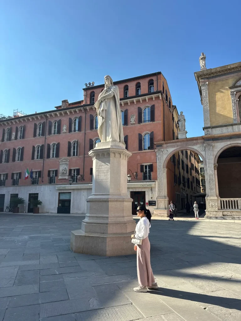 A woman standing in Piazza dei Signori in Verona beside the statue of Dante Alighieri, with historic buildings and an arched passage in the background under a clear blue sky.