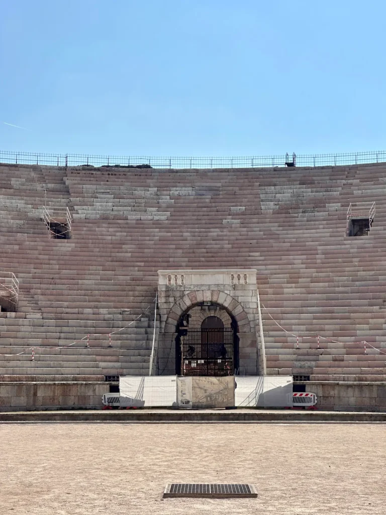 Interior of the Arena di Verona showing empty stone seating tiers and central archway under a clear blue sky