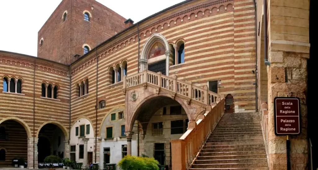Historic courtyard view of Palazzo della Ragione in Verona, Italy, featuring striped brickwork, arched windows, and the grand Scala della Ragione staircase leading up to a balcony.