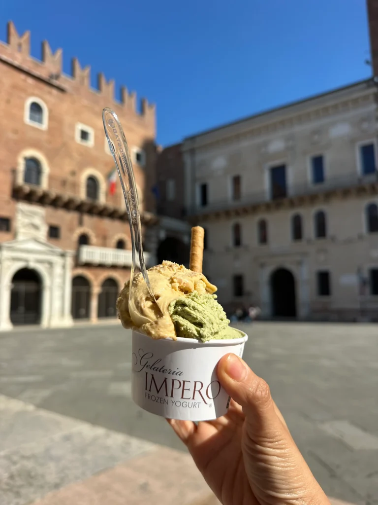 A close-up of a cup of pistachio and tiramisu gelato fro Gelato Impero with a cookie stick and spoon, held up in front of historic buildings in Verona’s Piazza dei Signori.
