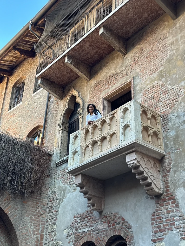 Me standing smiling on Juliet’s iconic stone balcony, which juts from the aged brick facade of Casa di Giulietta in Verona, beneath a clear blue sky.