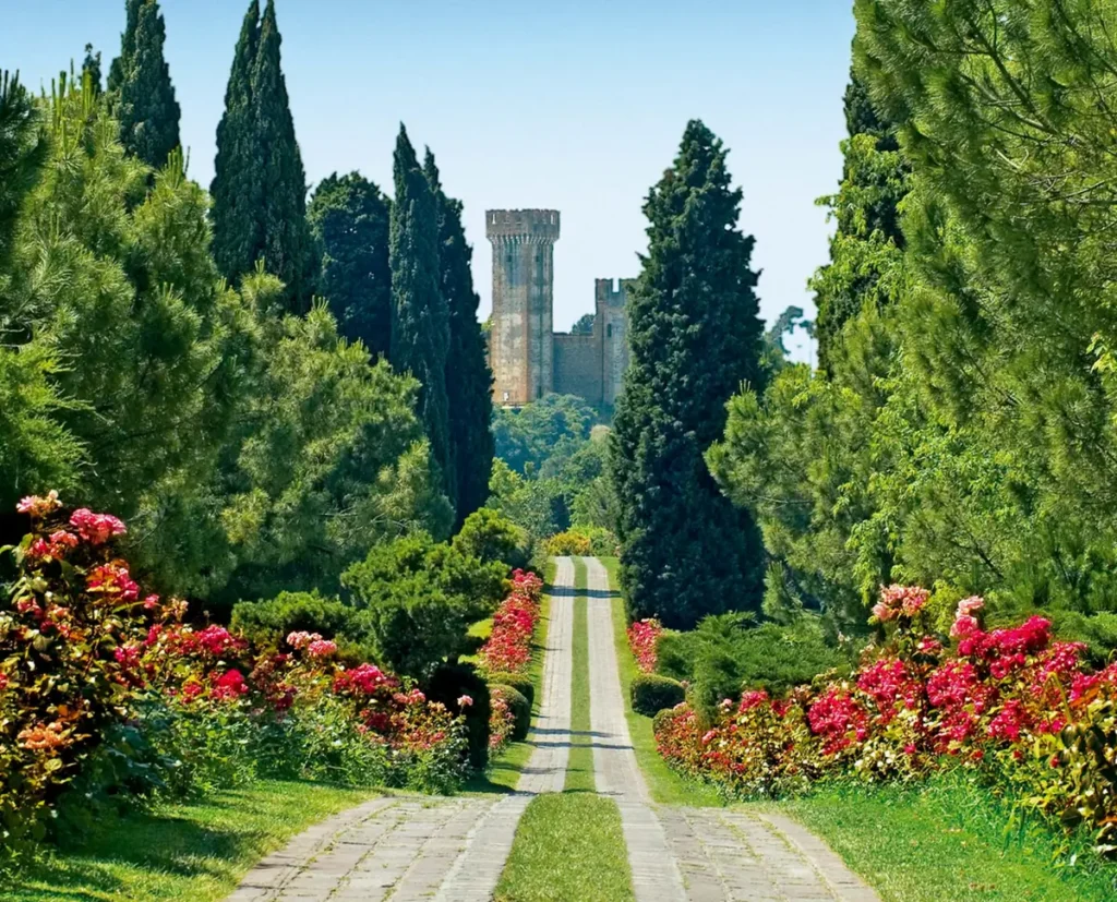 A picturesque pathway at Parco Giardino Sigurtà near Verona, Italy, lined with blooming pink and red flowers and tall, lush cypress and pine trees. In the distance, a medieval stone tower rises above the treetops.