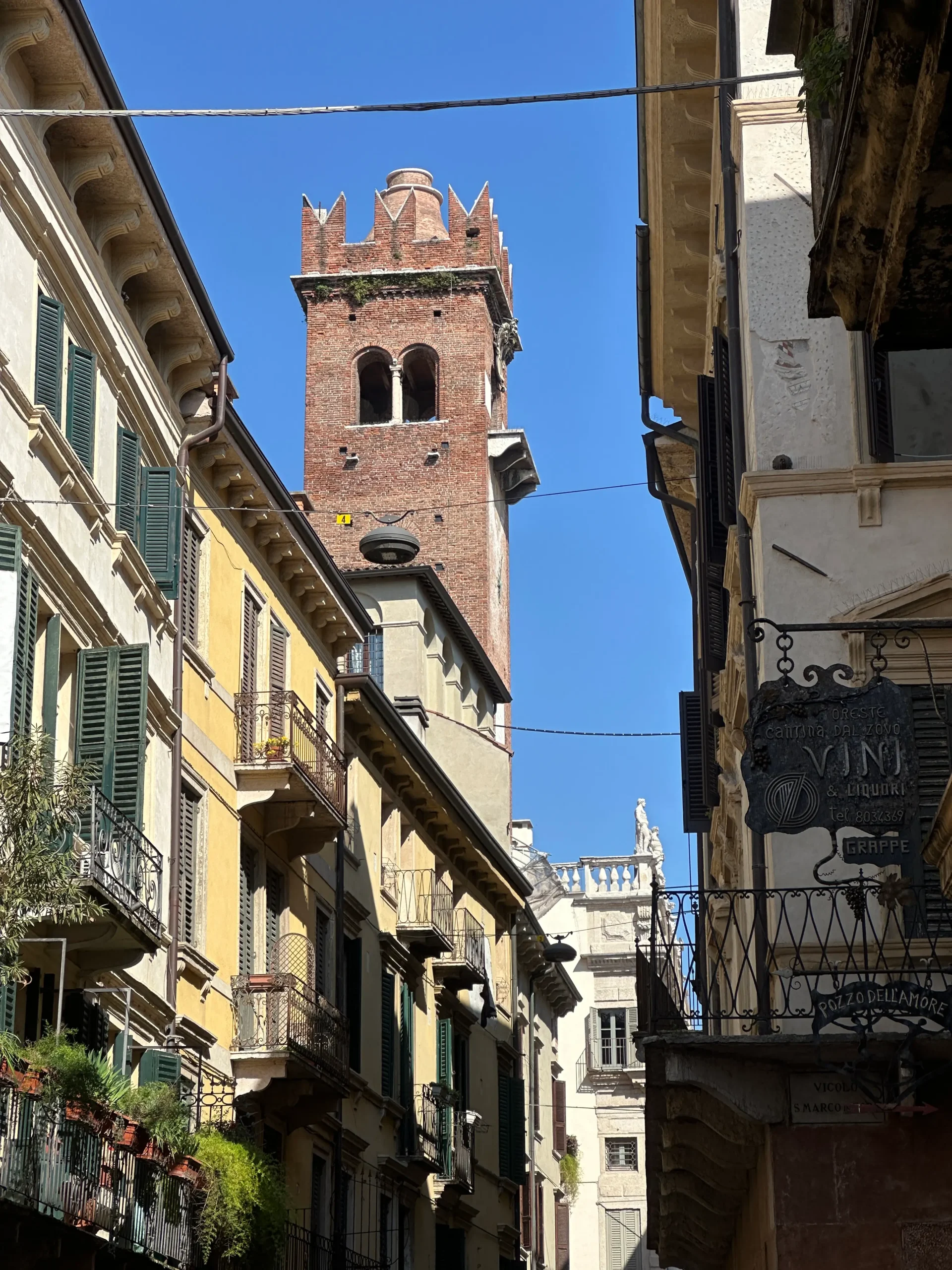 Narrow street in Verona’s historic center with tall, colorful buildings featuring green shutters and wrought iron balconies, leading to a medieval red-brick bell tower under a clear blue sky.
