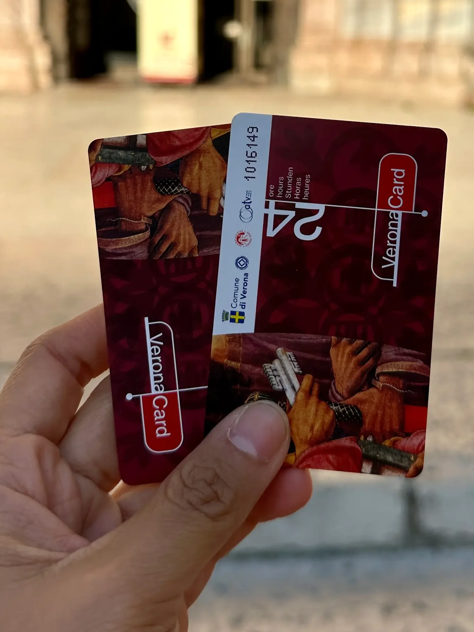 IA hand holds two red Verona Cards in front of the entrance to Verona Cathedral. A contemplation for "Is Verona Card worth it"