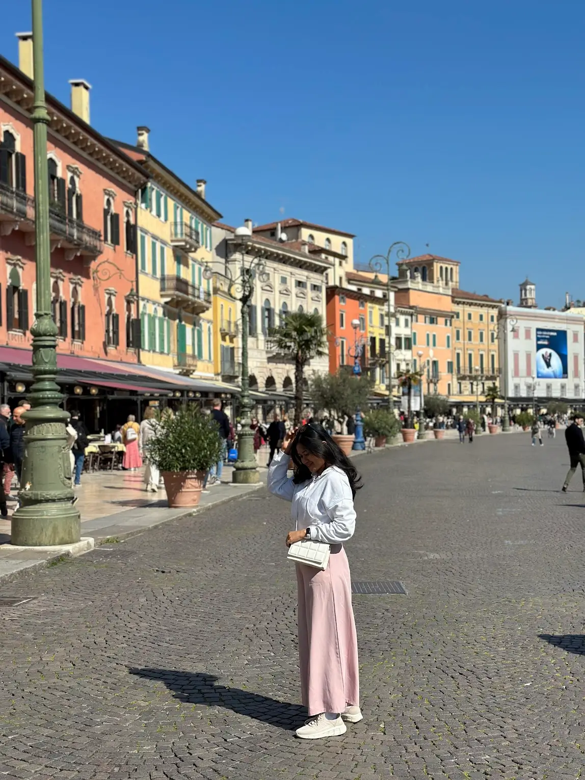 Person standing in Piazza Bra, Verona, with colorful historic buildings lining the square and the Arena area visible nearby on a sunny day.