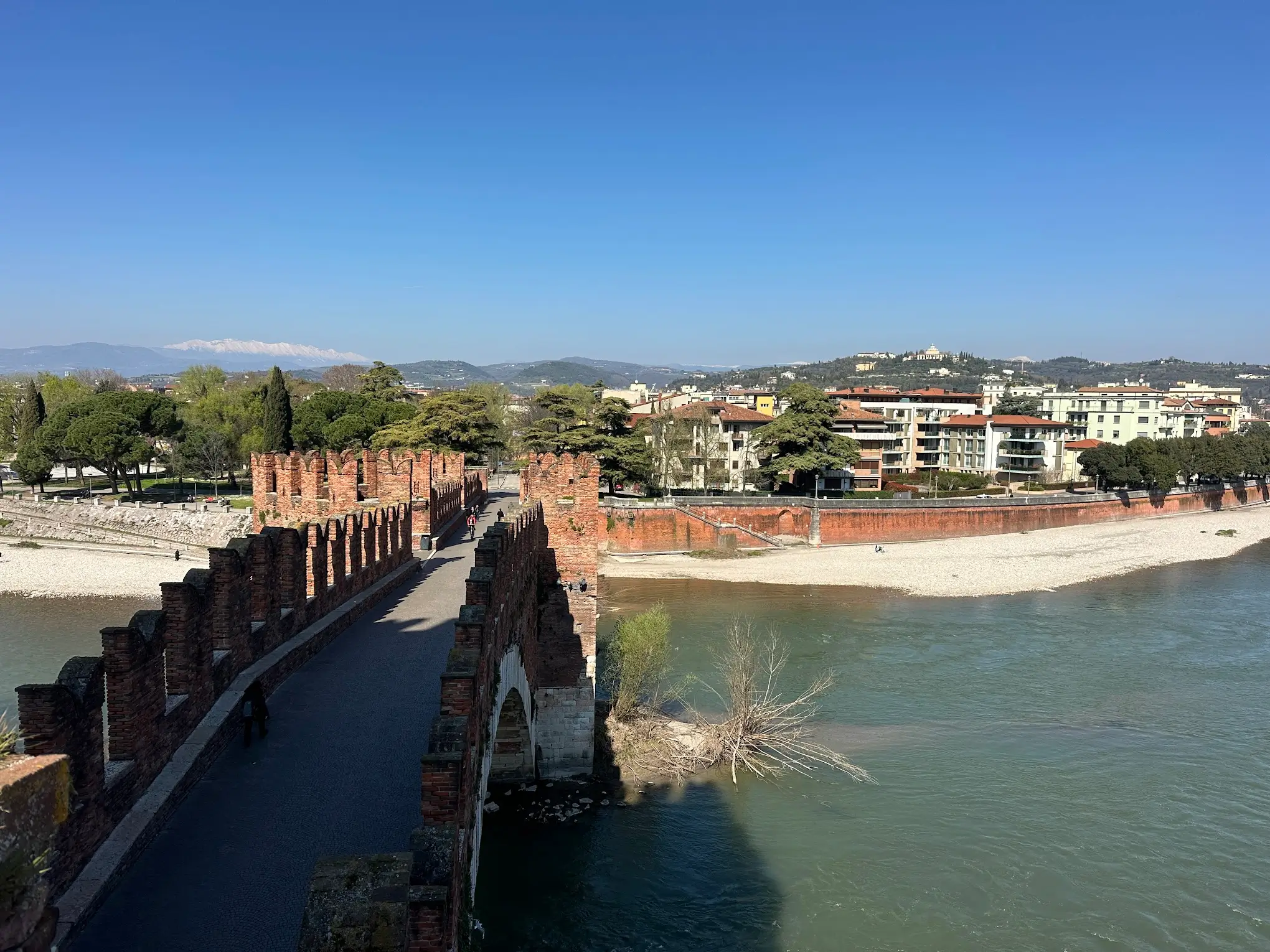 View from Ponte Scaligero in Verona, showing the brick medieval bridge stretching across the Adige River, with calm green water, red city walls, and residential buildings under a clear blue sky.

