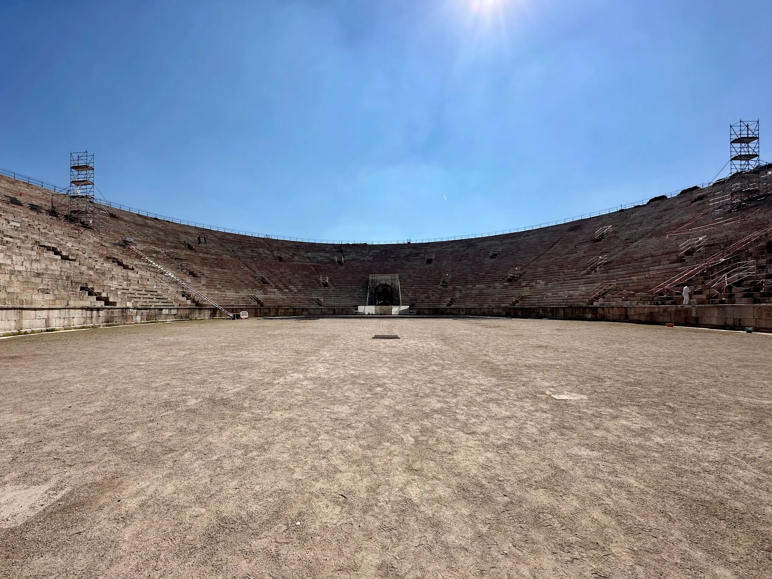 Wide view of the arena floor and stone seating inside the Arena di Verona amphitheater during a daytime visit, with renovation scaffolding visible on the upper tiers.