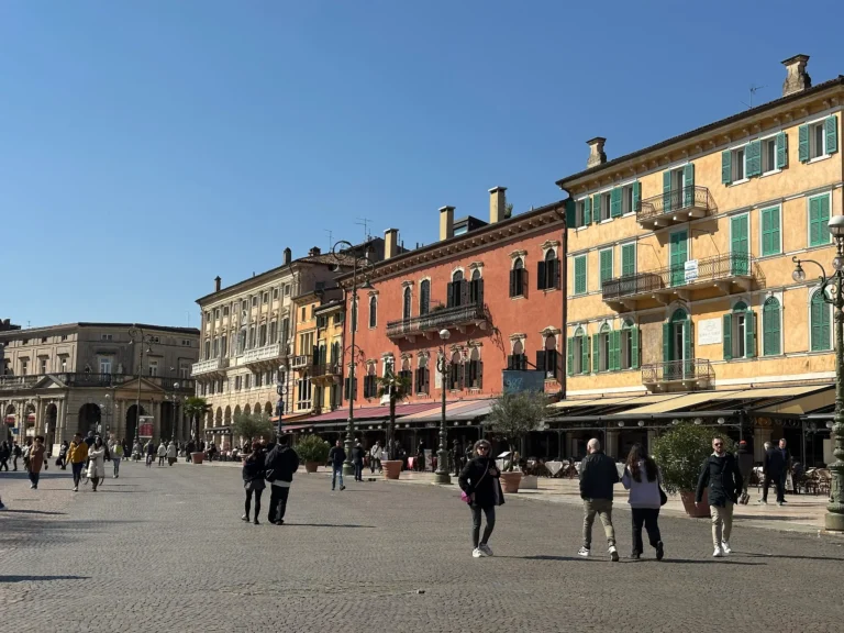 Wide view of Piazza Bra in Verona with colorful historic buildings, outdoor cafes, and people walking across the open square under a clear blue sky.