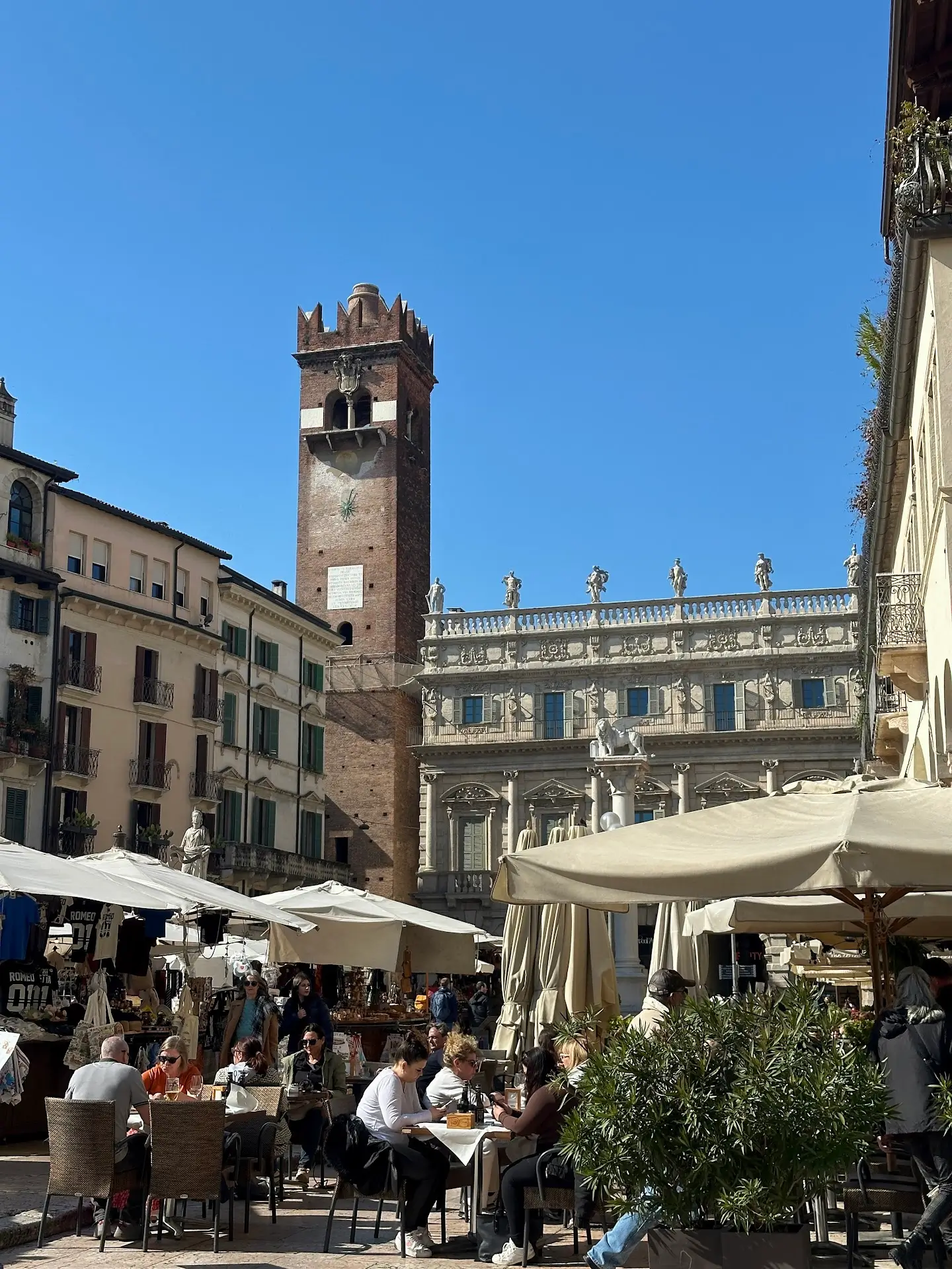 Where to eat in Verona at Piazza delle Erbe, with outdoor dining, market stalls, and historic buildings under a clear blue sky.