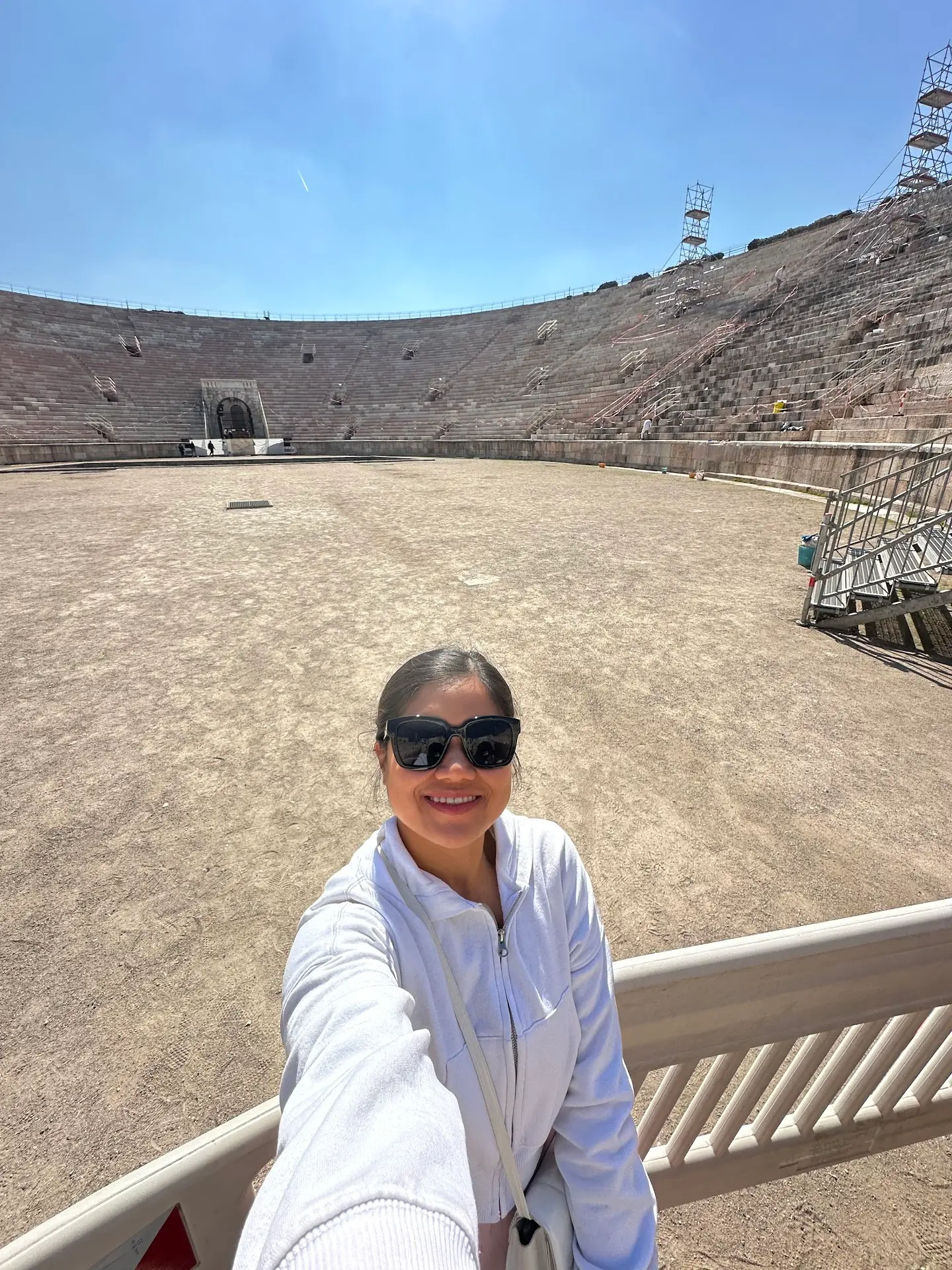 Person taking a selfie inside the Arena di Verona amphitheater during daytime, with the large stone seating and arena floor visible behind, and renovation scaffolding on the side.