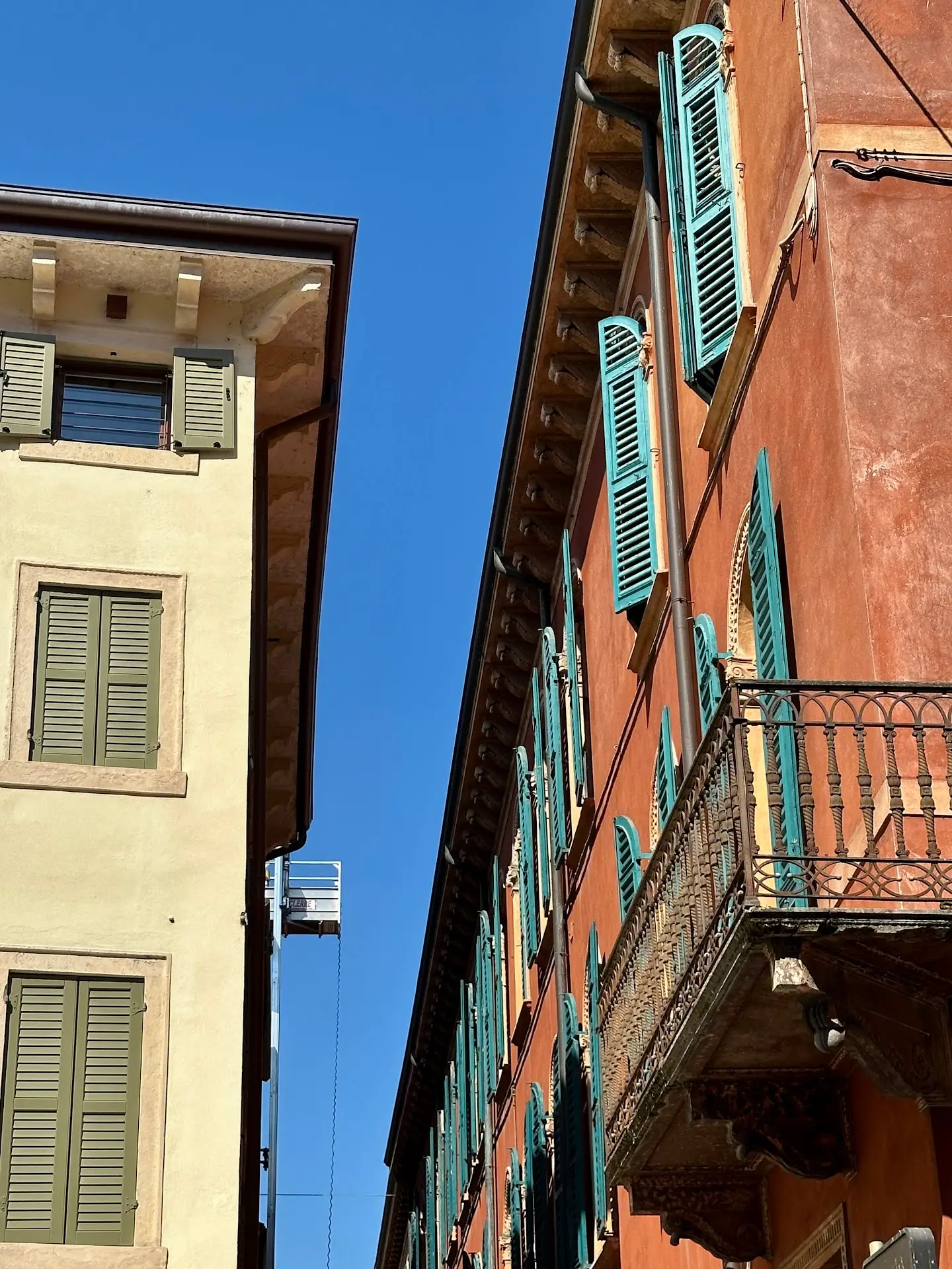Close-up of traditional Italian buildings with pastel walls, green shutters, and a small wrought-iron balcony against a bright blue sky.