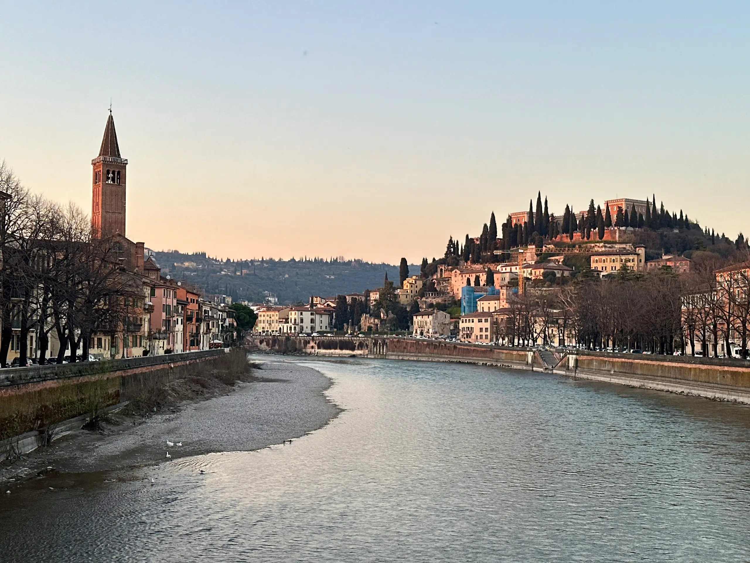 View of the Adige River in Verona at sunset, with Ponte Pietra area in the distance, historic buildings along the riverbanks, and Castel San Pietro on the hill.