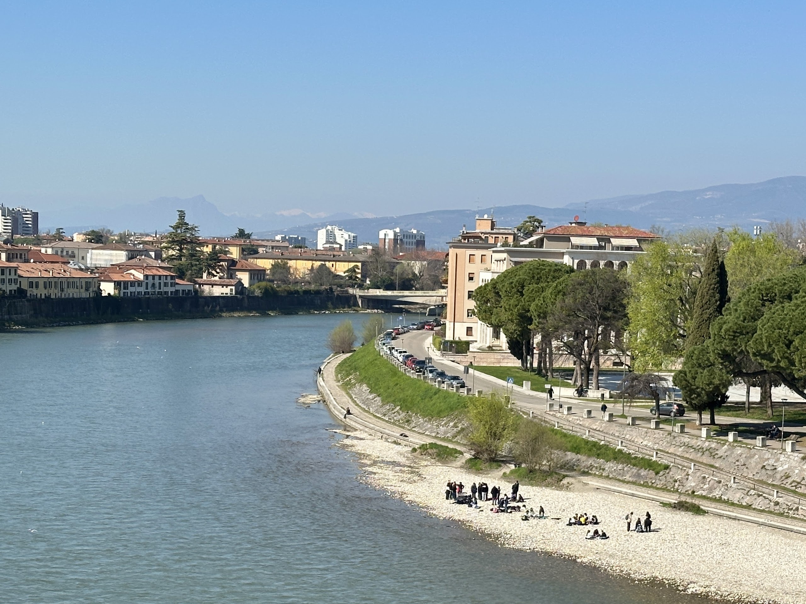View of the Adige River in Verona with a riverside walkway, people relaxing on the bank, and buildings lining the opposite shore