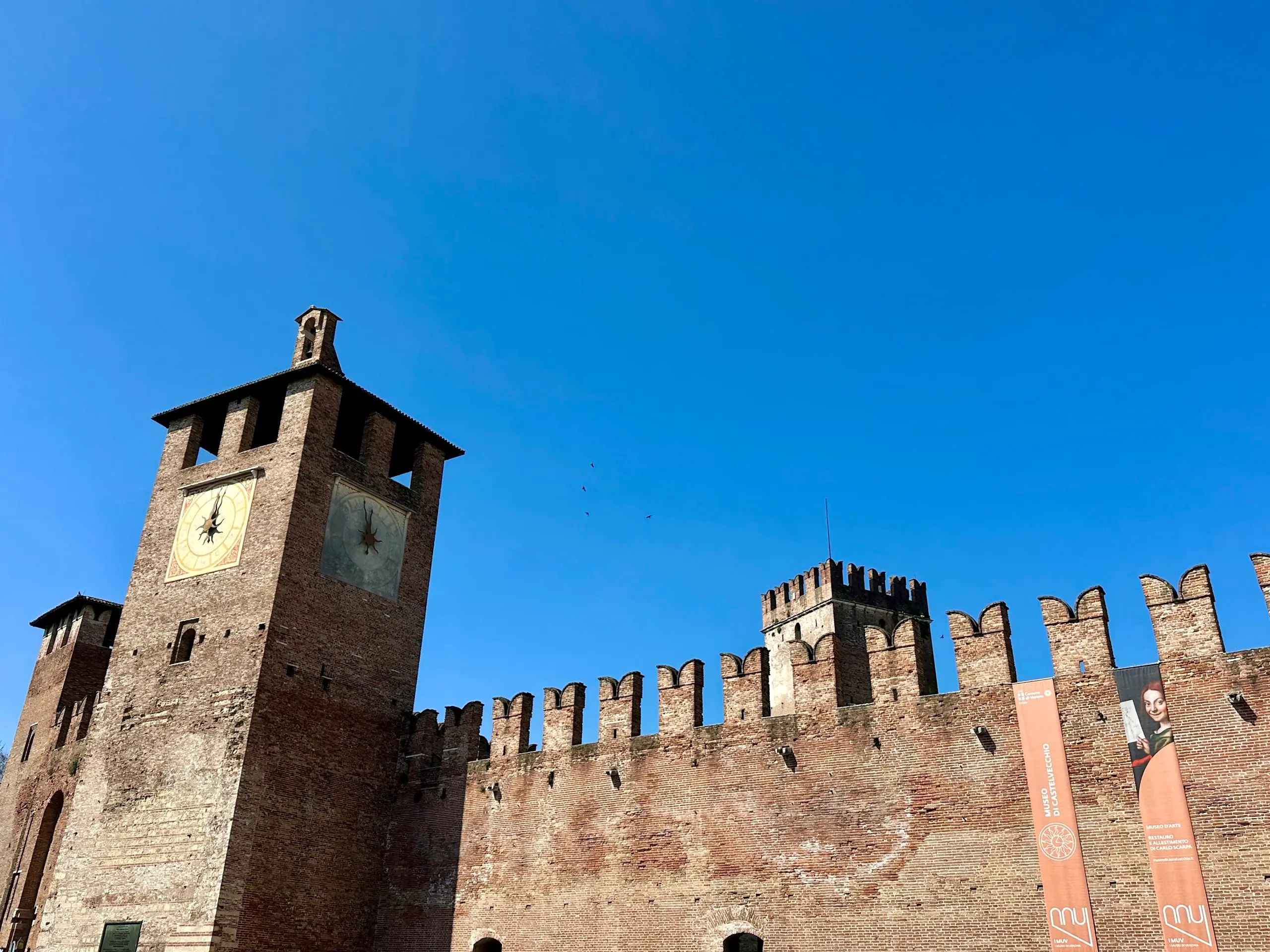Castelvecchio Verona fortress wall with crenellations and a square clock tower under a clear blue sky.